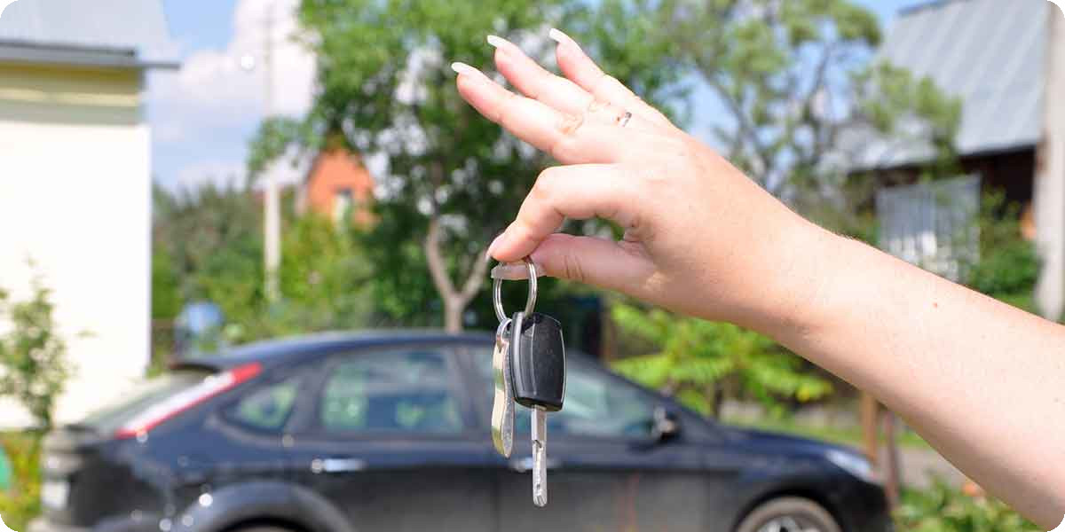 Eine Frau hält einen Autoschlüssel in die Kamera und im Hintergrund steht ein schwarzes Auto.