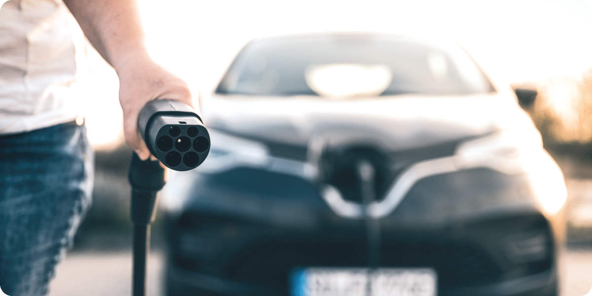 Frau steht vor einen Elektroauto und hält den Stecker vom Ladegerät in der Hand.