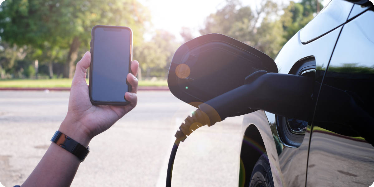 Person hält Smartphone in Kamera und lädt Elektroauto im Hintergrund.